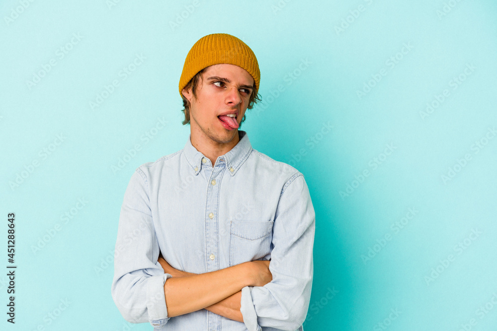 Young caucasian man with make up isolated on blue background  funny and friendly sticking out tongue.