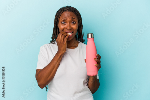 Young african american woman holding canteen isolated on blue background biting fingernails, nervous and very anxious.