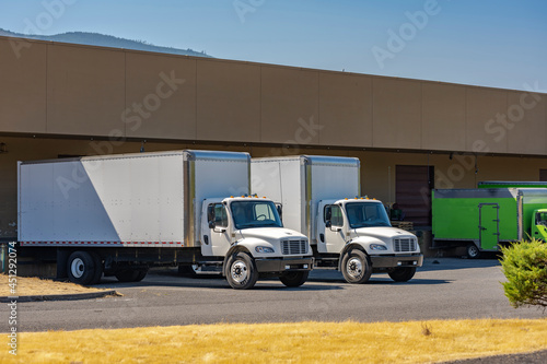 Compact medium size rigs day cab semi trucks with long box trailers for local delivery standing at warehouse dock loading commercial cargo