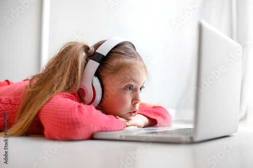 Cute little girl in headphones seriously using laptop at home for online education, home studying and online communication. Kids distance learning. Social distance. Stay at home. photo