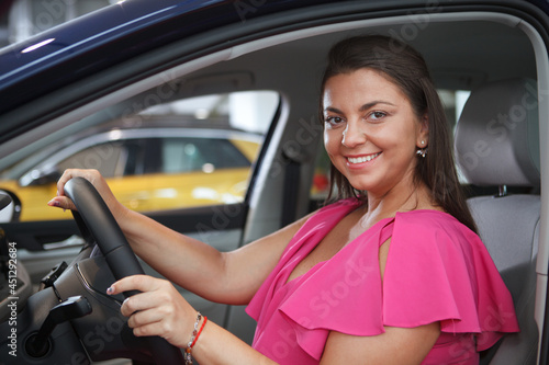 Charming happy woman smiling to the camera while sitting in drivers seat of a car, holding steering wheel