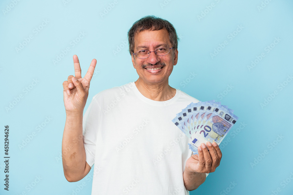 Senior indian man holding bill isolated on blue background showing number two with fingers. Senior indian man holding bill isolated on blue background showing number two with fingers.