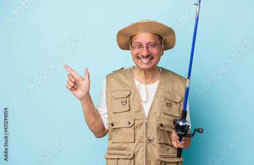 Senior indian fisherman holding rod isolated on blue background smiling and pointing aside, showing something at blank space.