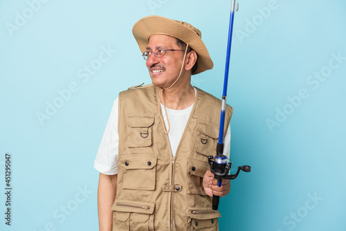 Senior indian fisherman holding rod isolated on blue background looks aside smiling, cheerful and pleasant.