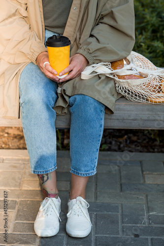 A modern stylish woman with a paper cup of hot coffee in her hands and a string bag with fresh bread on a bench. The concept of street food and ecology. Refusal of plastic bags.