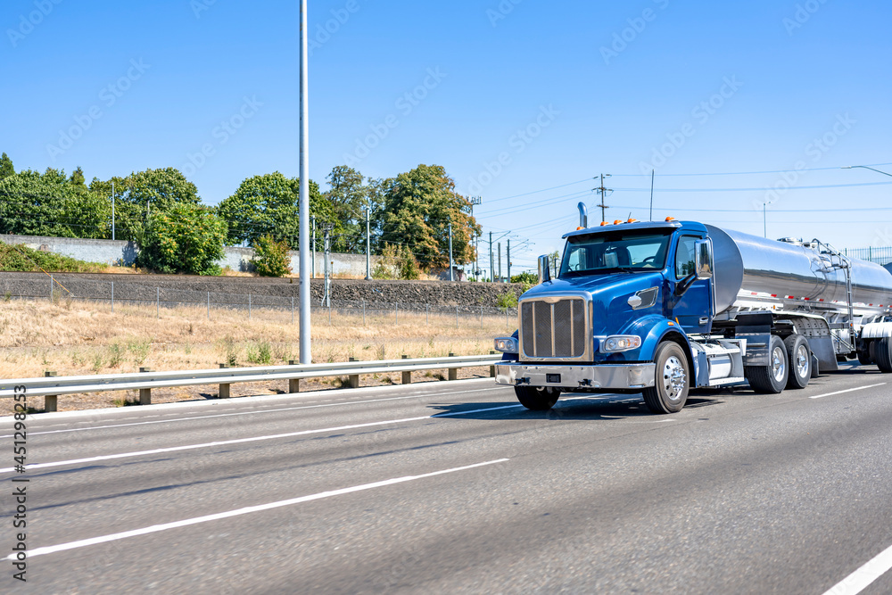 Powerful blue day cab big rig semi truck transporting liquid cargo in shiny tank semi trailer driving on the straight multiline highway road