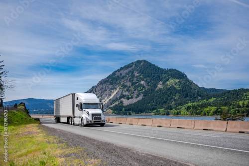 Powerful industrial white big rig semi truck with grille guard transporting frozen cargo in reefer semi trailer driving on highway road with river and mountain on the side