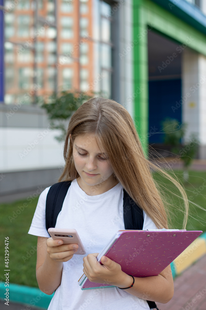 Obraz premium Cute schoolgirl on the street with a smartphone, notebooks, and a backpack. Back to school. Concept education