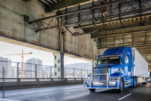 Stylish blue big rig industrial grade semi truck with grille guard transporting cargo in refrigerator semi trailer running on the two level Fremont bridge across the Willamette River
