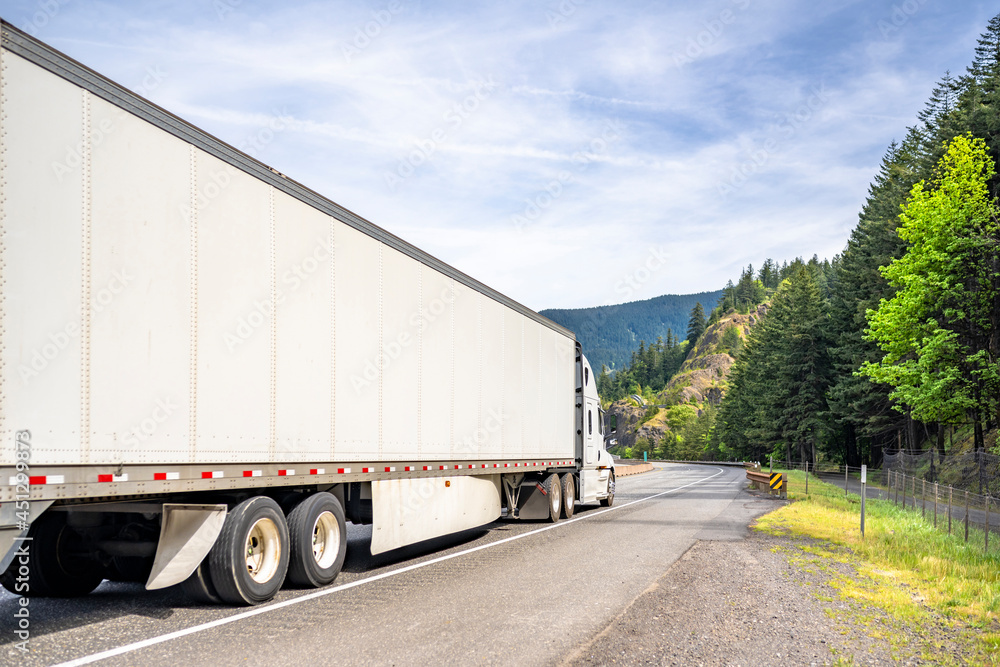 White big rig long haul industrial semi truck transporting goods in dry van semi trailer with skirt driving on the winding road with mountain on the side