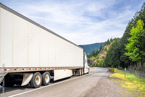 White big rig long haul industrial semi truck transporting goods in dry van semi trailer with skirt driving on the winding road with mountain on the side