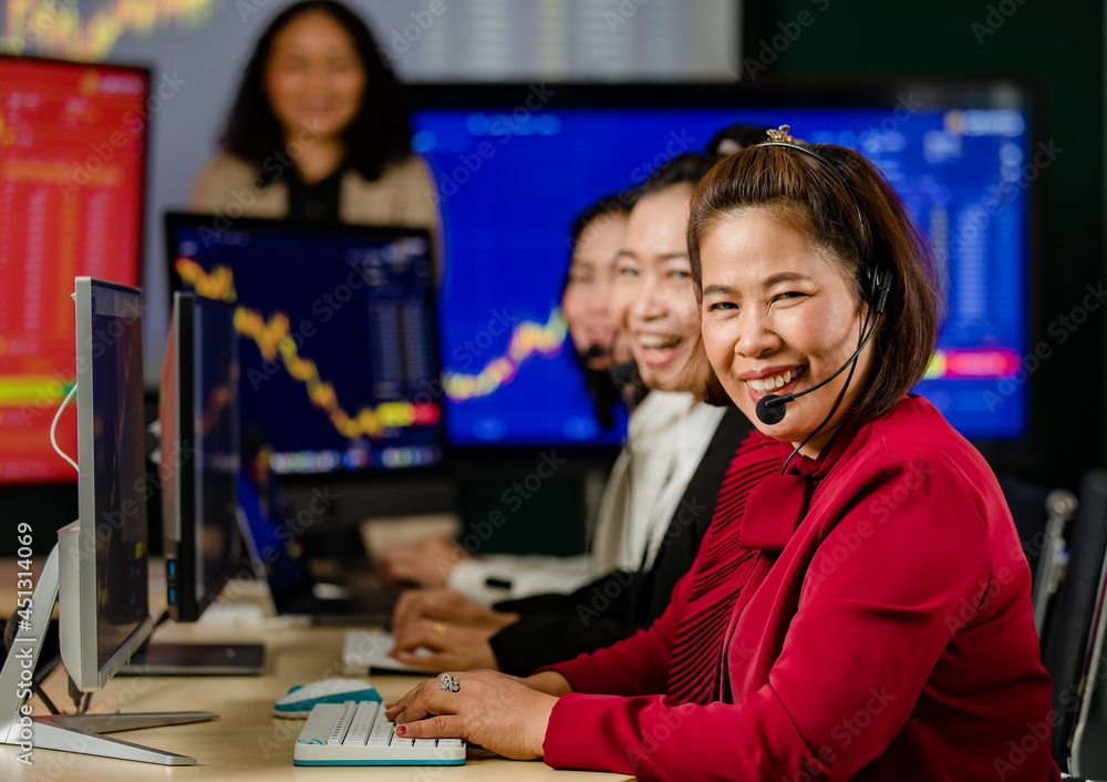 Fototapeta premium Group of Asian female customer service operators wears microphone headsets sit smiling in front daily graph chart computer monitor screen talking and trading buy sell stock market online with client