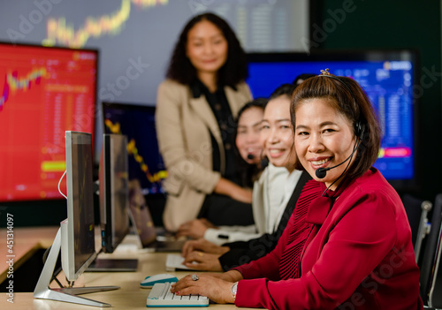 Asian female professional stock exchange manager sit on desk smile in front chart monitor look at camera while customer service operator team wears microphone headsets working in blurred foreground