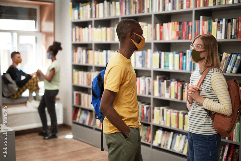 Side view portrait of two students wearing masks while chatting in school library, copy space