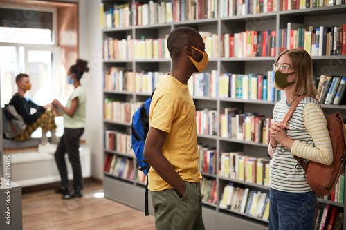 Side view portrait of two students wearing masks while chatting in school library, copy space
