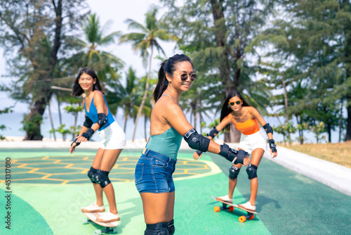 Group of Happy Asian woman friends skateboarding together at skateboard park by the beach. Happy female friendship enjoy summer outdoor active lifestyle play extreme sport surf skate at public park.