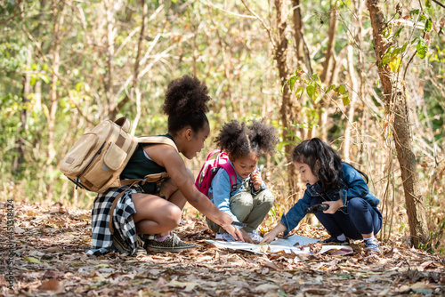 Group of Diversity little girl friends with backpack hiking together at forest mountain in summer sunny day. Three kids having fun outdoor activity sitting and looking at the map exploring the forest. photo