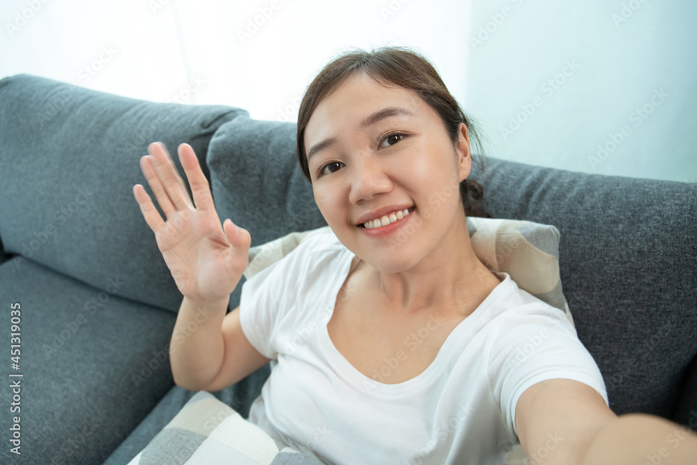 Beautiful Asian woman vlogger motion her hand and smile to camera while making a video call. Young woman relaxing in living room and rising her hand while using video call to her family. Beautiful Asian woman vlogger motion her hand and smile to camera while making a video call. Young woman relaxing in living room and rising her hand while using video call to her family.