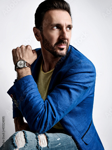 Portrait of mature bearded man, businessman in modern torn jeans and blue jacket sitting on stool sideways and looking behind him over white background