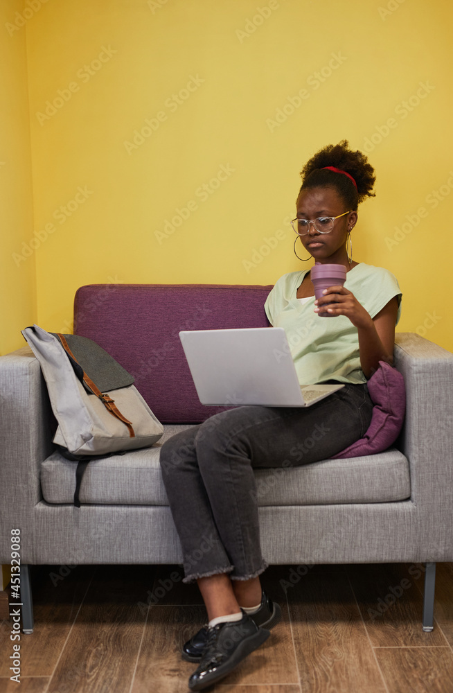 Vertical full length portrait of young African-American woman using laptop while relaxing at college campus, copy space Vertical full length portrait of young African-American woman using laptop while relaxing at college campus, copy space
