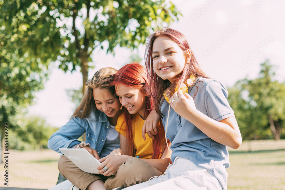 Fototapeta premium Attractive teenage girls are sitting on a park bench, discussing and smiling. Girlfriends are chatting in the park