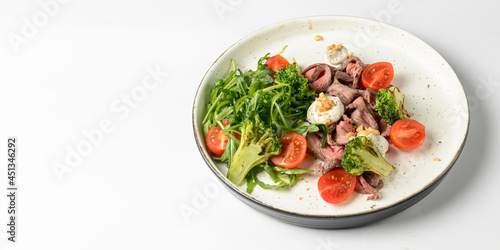 Healthy salad made from meat, cherry tomatoes, broccoli, goat cheese and fresh arugula served in a white plate.