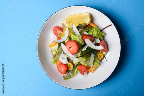 Spinach salad with strawberries, cherry tomatoes and coconut chip slices in a white plate over pastel blue background.