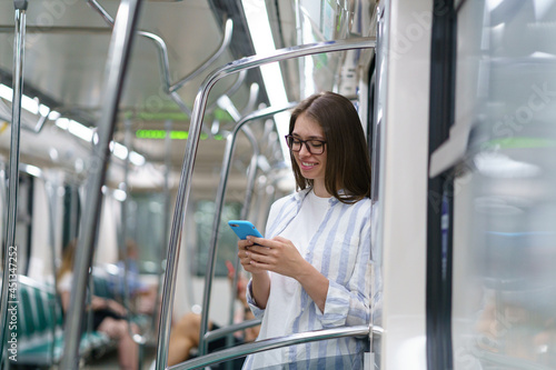 Young passenger woman texting message on cellphone, chatting in social networks. Smiling millennial girl using mobile smart phone in subway train at metro. Public transportation, underground. 
