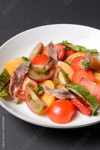 Vegetable salad made of tomatoes and fish slices, served in a white plate over black background.