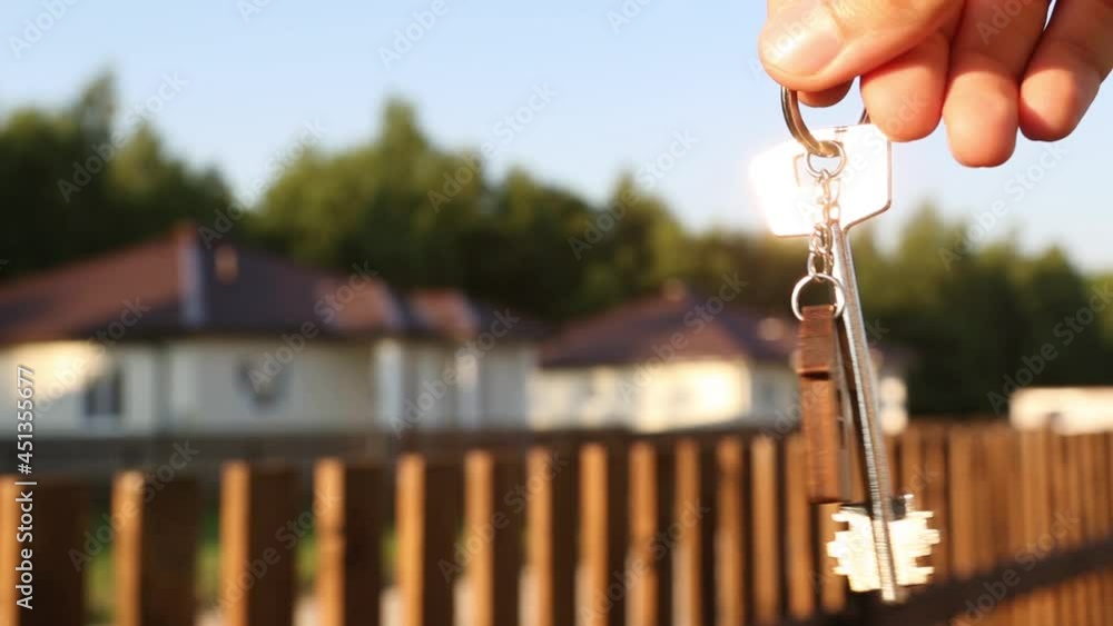 Key ring with keychain house in hand. Background of fence and cottage. Moving to a new home, mortgage, buying real estate, renting and booking housing, dream of living in the village. Copy space Key ring with keychain house in hand. Background of fence and cottage. Moving to a new home, mortgage, buying real estate, renting and booking housing, dream of living in the village. Copy space