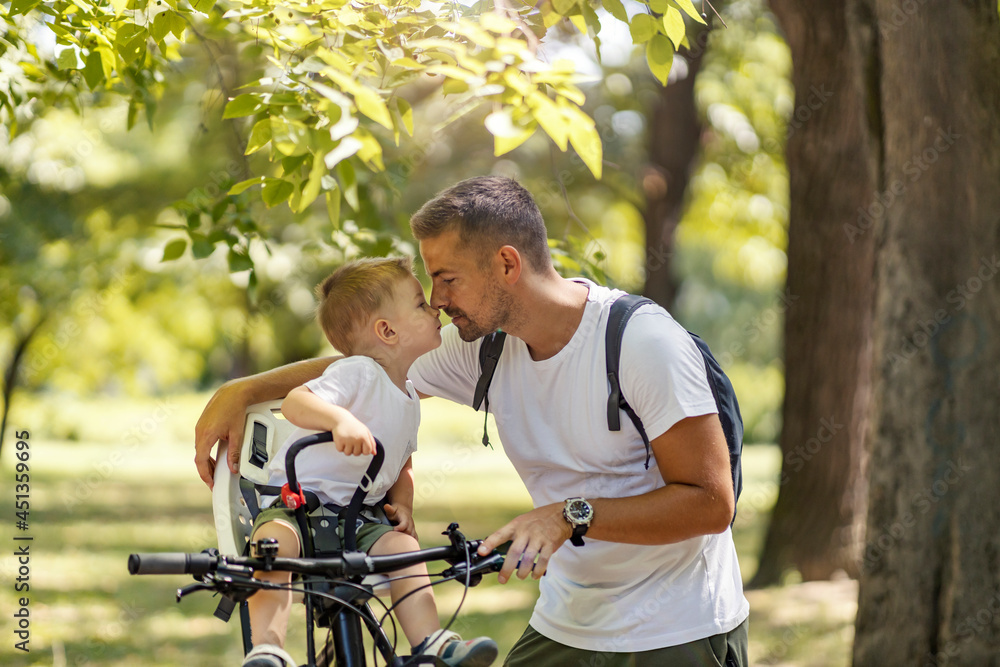 Emotional family moment. Father and son ride a bike through the woods on a sunny summer day. A cute boy is sitting in a bicycle basket and kissing his dad who is facing him. Family weekend in nature