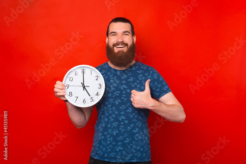 Pleased bearded man holding clock while showing thumb up and looking at the camera over red background
