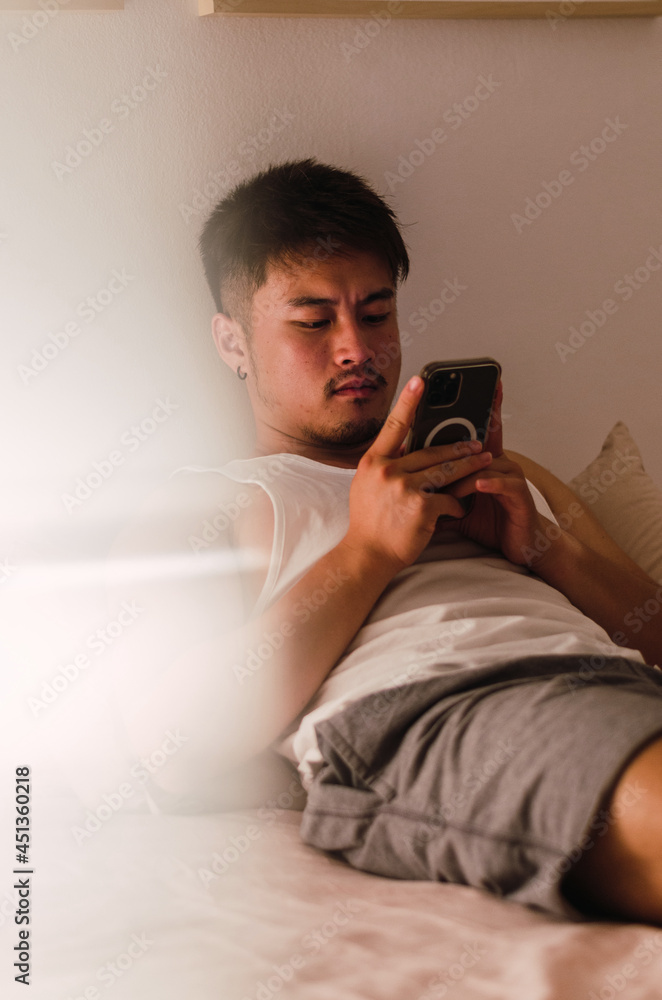 Fototapeta premium Young handsome Chinese man at home lying in bed using smartphone