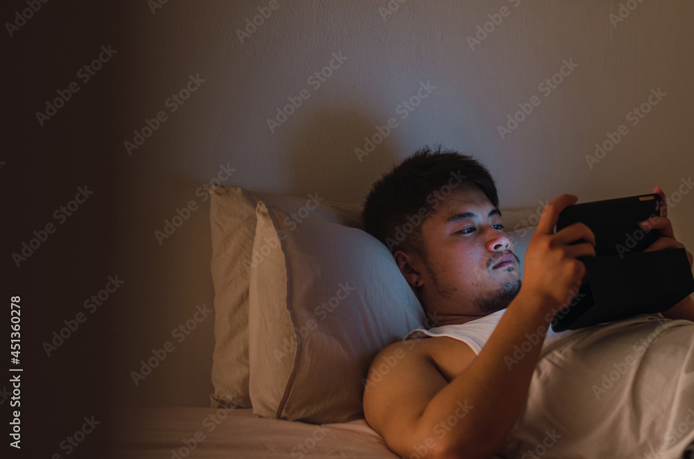 Fototapeta premium Young handsome Chinese man at home lying in bed using digital tablet