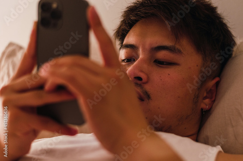 Young handsome Chinese man at home lying in bed using smartphone
