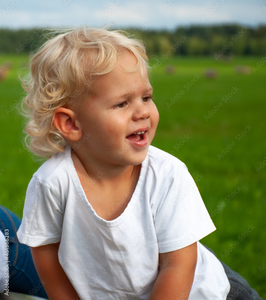 Little blonde curly-haired boy in a white T-shirt, laughing against the background of blue sky and green grass Little blonde curly-haired boy in a white T-shirt, laughing against the background of blue sky and green grass