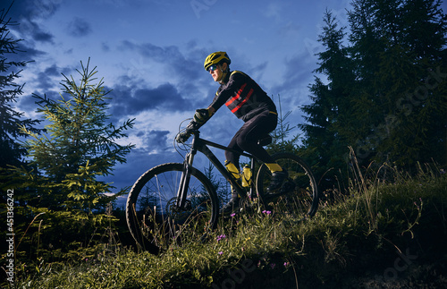 Young man riding bicycle downhill with beautiful blue evening sky on background. Male bicyclist in sports cycling suit cycling down grassy hill at night. Concept of sport, biking and active leisure.