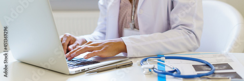 Cropped photo of female doctor in white uniform working on laptop computer in hospital, sitting at desk with stetoscope and ultrasound results. Medical panoramic banner for website header