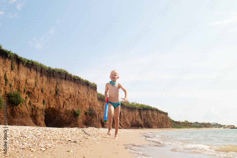 A boy runs along the beach in swimming trunks. He has a water pistol in his hands. Happy child on the sandy beach. Concept of happy life and relaxation by the ocean. Blond boy smiling at the camera
