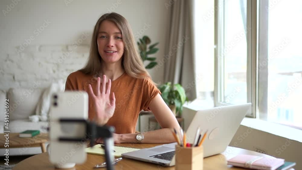 Woman talking and waving in front of phone screen while sitting at table in home office. Woman talking and waving in front of phone screen while sitting at table in home office.
