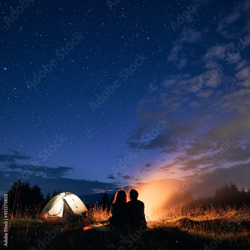 Beautiful view of night starry sky over grassy hill with illuminated camp tent  campfire and hikers. Back view of couple tourists having a rest near bonfire. Concept of night camping  relationship.