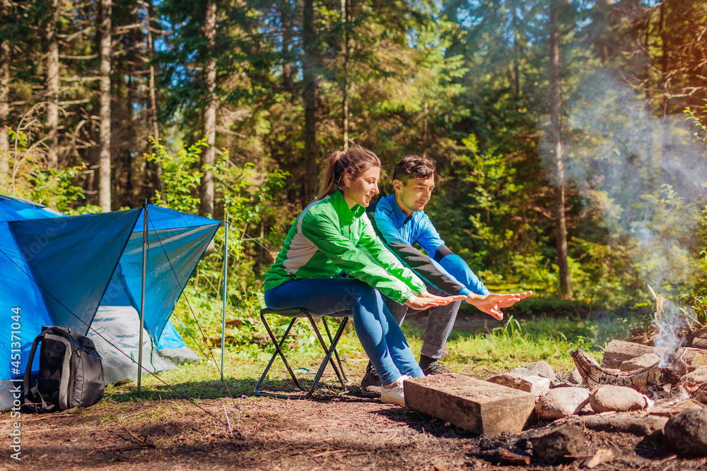 Young couple of travelers enjoy campfire in summer forest. Tourists sitting by tent warming hands. Camping in woods Young couple of travelers enjoy campfire in summer forest. Tourists sitting by tent warming hands. Camping in woods