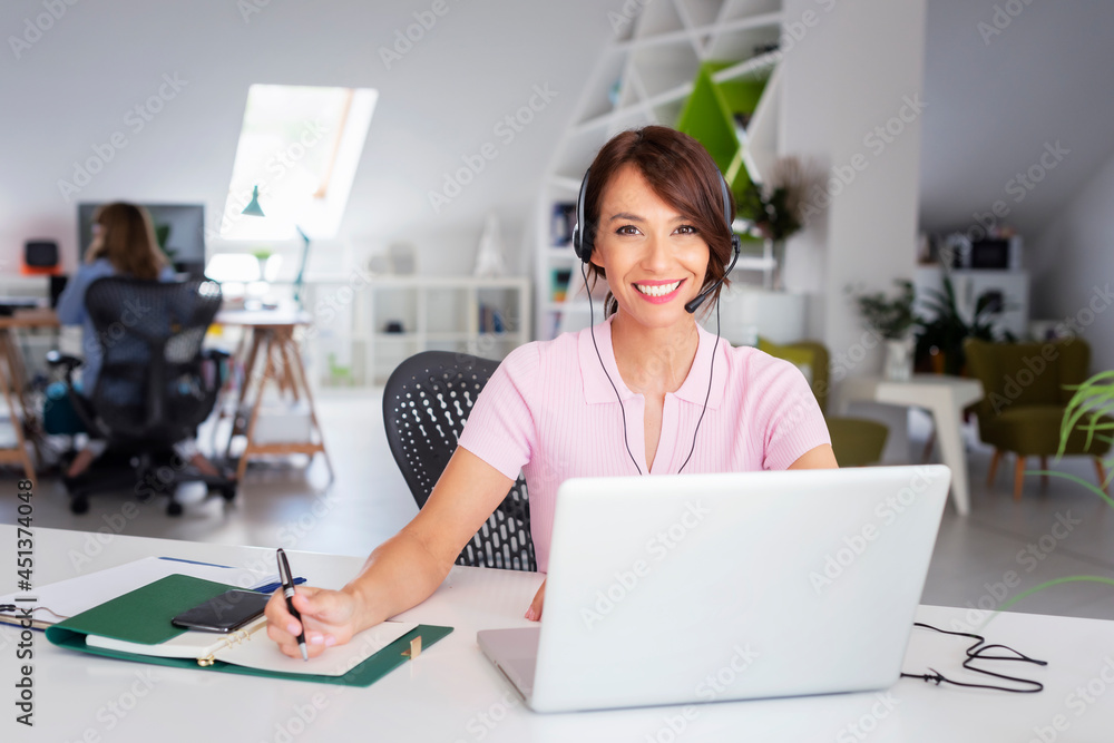 Attractive businesswoman having online meeting while working at the office. Customer service assistant wearing headset while sitting behind her computer and working in the call center. Attractive businesswoman having online meeting while working at the office. Customer service assistant wearing headset while sitting behind her computer and working in the call center.