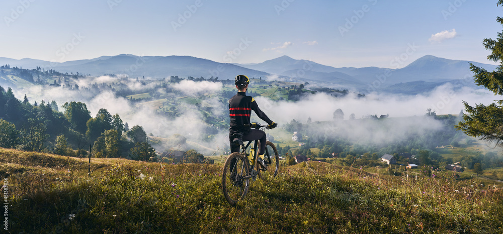 Man riding bicycle on grassy hill and looking at beautiful misty mountains. Male bicyclist enjoying panoramic view of majestic mountains during bicycle ride. Concept of sport, bicycling and nature.