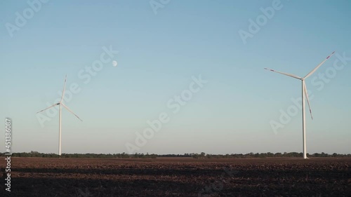 a lonely wind turbine stands in an unseeded field. the moon is visible in the sky, summer sunset photo