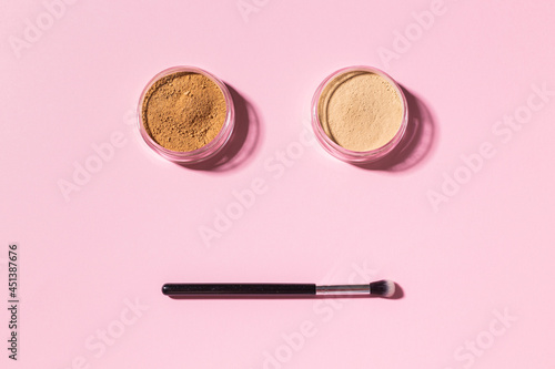 Funny face with make-up brushes and mineral powder on pink background with copy space, top view. Cosmetics humor and beauty concept.