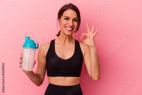 Young caucasian woman drinking a protein shake isolated on pink background cheerful and confident showing ok gesture.