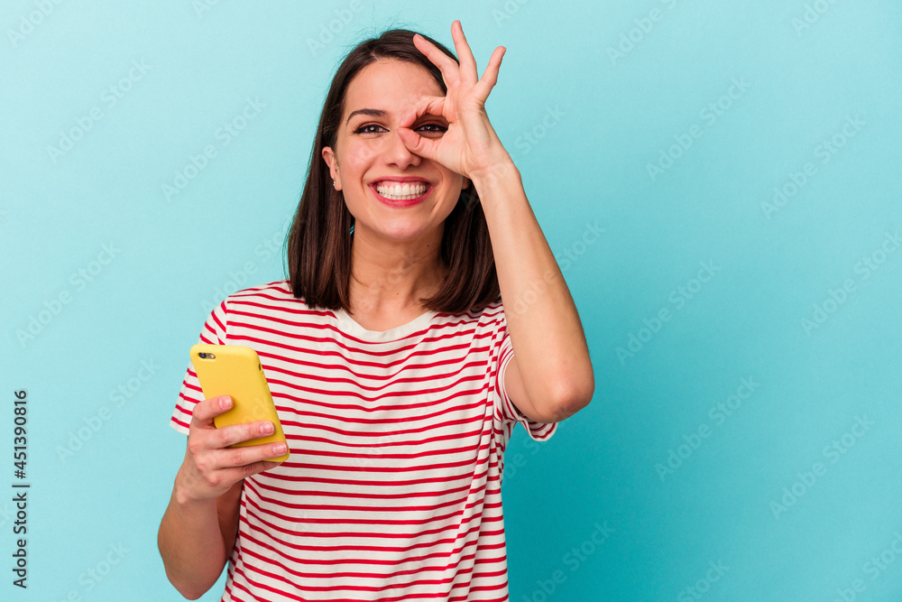 Young caucasian woman holding mobile phone isolated on blue background excited keeping ok gesture on eye. Young caucasian woman holding mobile phone isolated on blue background excited keeping ok gesture on eye.