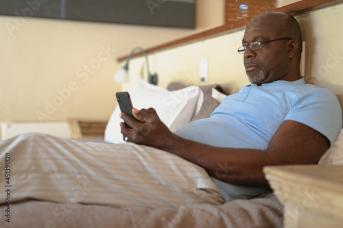 Senior african american man laying in the bad and using smartphone