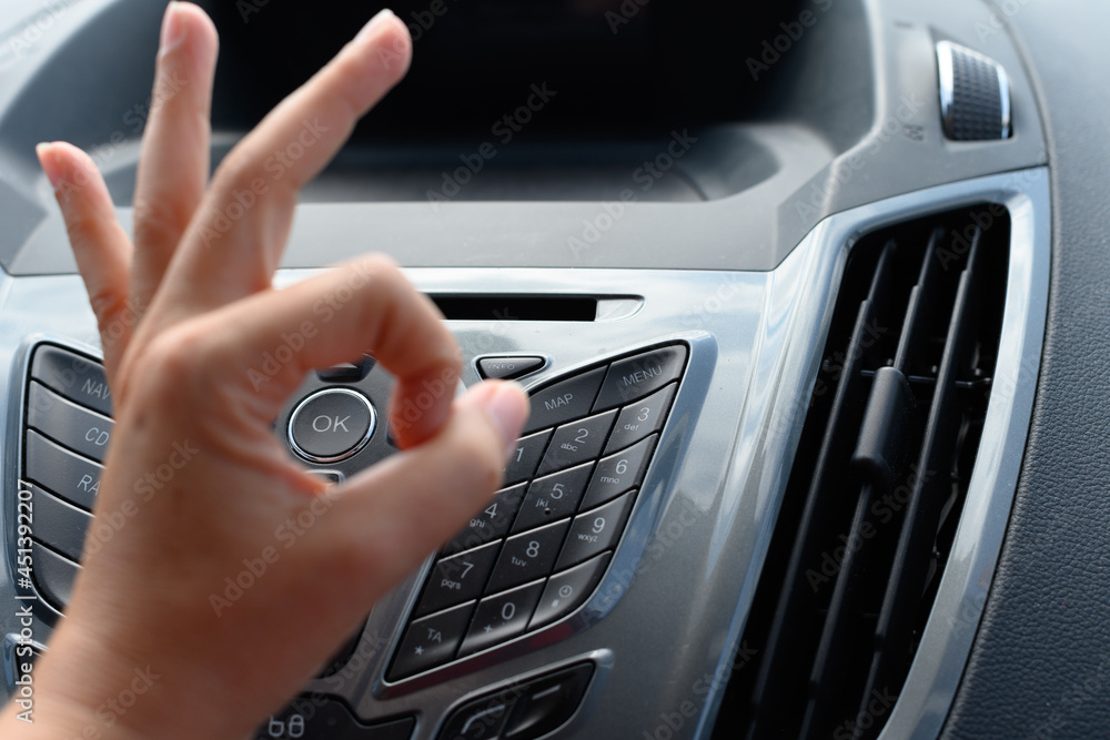 Panel of a car with a button with the ok sign and a hand in front with the ok sign as well.